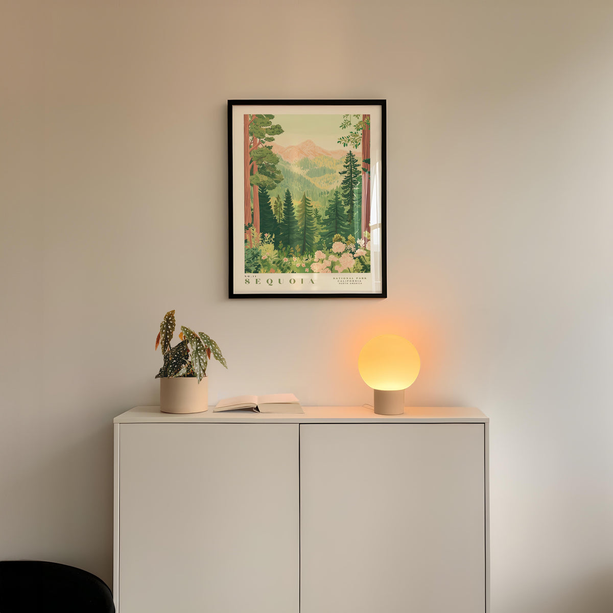 a framed poster of a forest scene hanging on a wall above a white cabinet with a plant and a lamp on top.