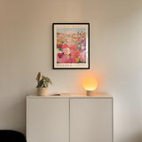 a framed poster of a colorful floral scene hanging on a wall above a white cabinet with a plant and a lamp on top.