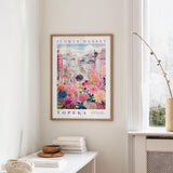 a framed poster of a colorful floral scene hanging on a wall, with a white table and a vase of flowers in the foreground.