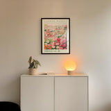 a white cabinet with a potted plant and a lamp on top, set against a white wall with a framed poster of a flower market above it.