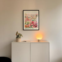a white cabinet with a potted plant and a lamp on top, set against a white wall with a framed poster of a flower market above it.