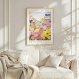 a framed poster of a colorful flower market scene hanging on a wall above a white couch with a blanket draped over it.