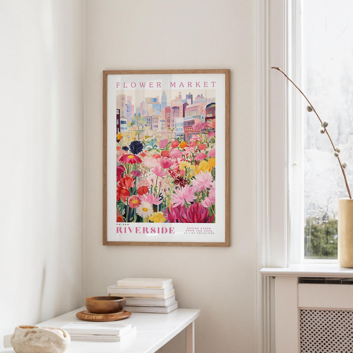 a framed poster of a colorful flower market scene hanging on a wall, with a white table and a vase of flowers in the foreground.