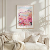 a framed poster of a flower market in Athens hanging on a wall above a white couch with a blanket draped over it.