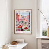 a framed poster of a colorful illustration of a flower market in New Orleans, with a cityscape in the background. The poster is hanging on a wall, and there is a small table with books and a vase nearby.