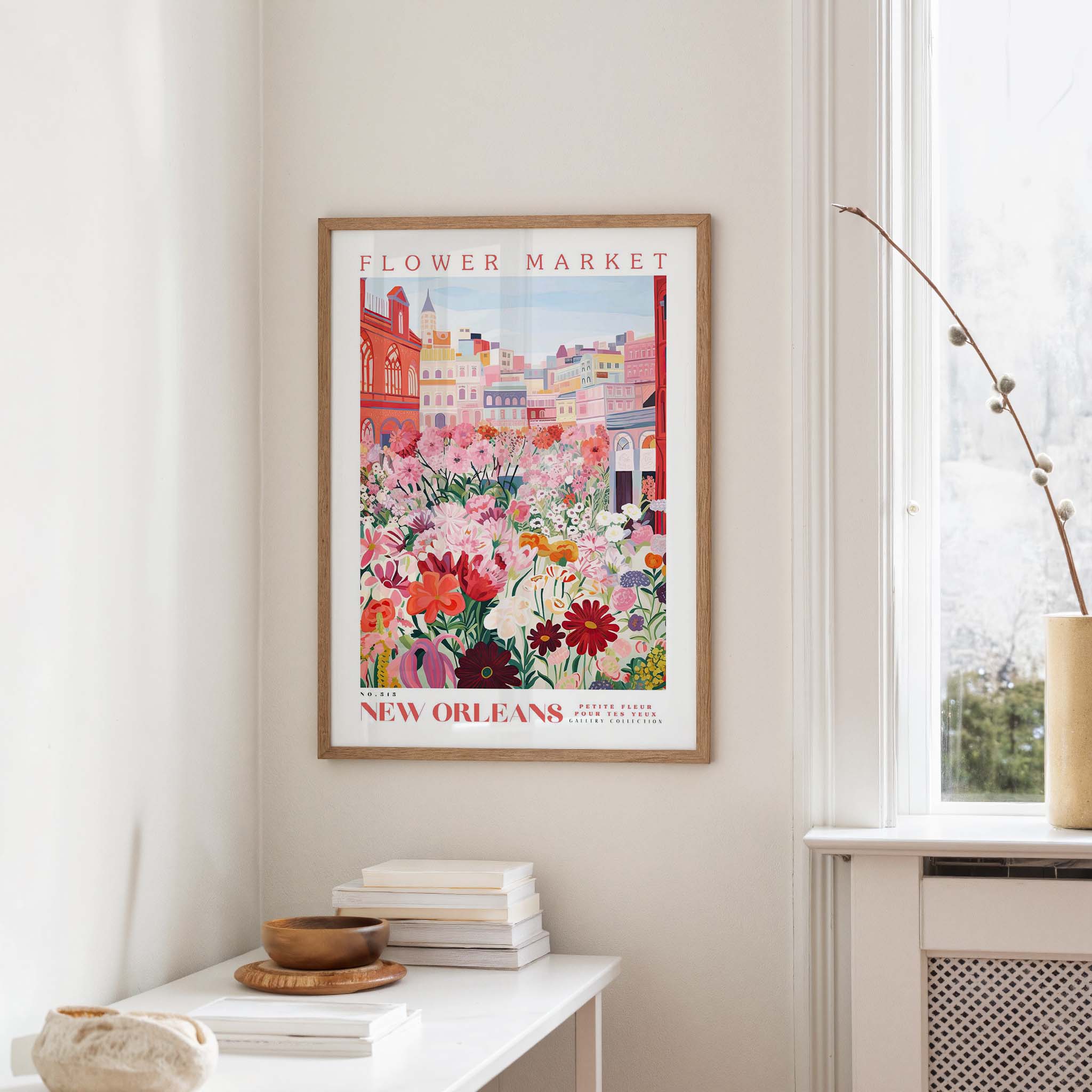 a framed poster of a colorful illustration of a flower market in New Orleans, with a cityscape in the background. The poster is hanging on a wall, and there is a small table with books and a vase nearby.