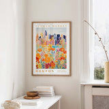 a framed poster of a colorful cityscape with flowers, hanging on a wall in a room with a white table and a window.