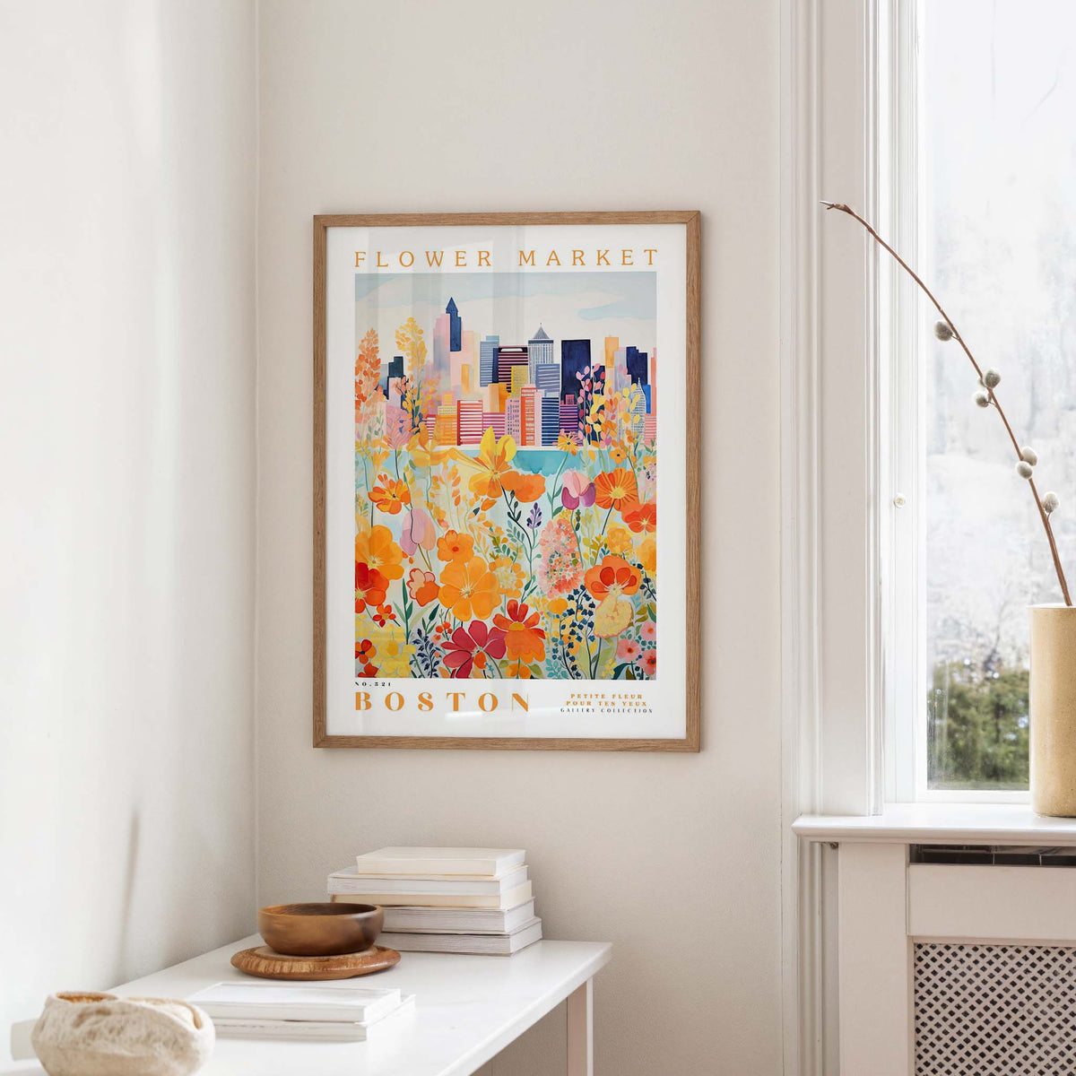 a framed poster of a colorful cityscape with flowers, hanging on a wall in a room with a white table and a window.