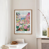 a framed poster of a colorful garden scene hanging on a wall, with a white table and a vase of flowers in the foreground.