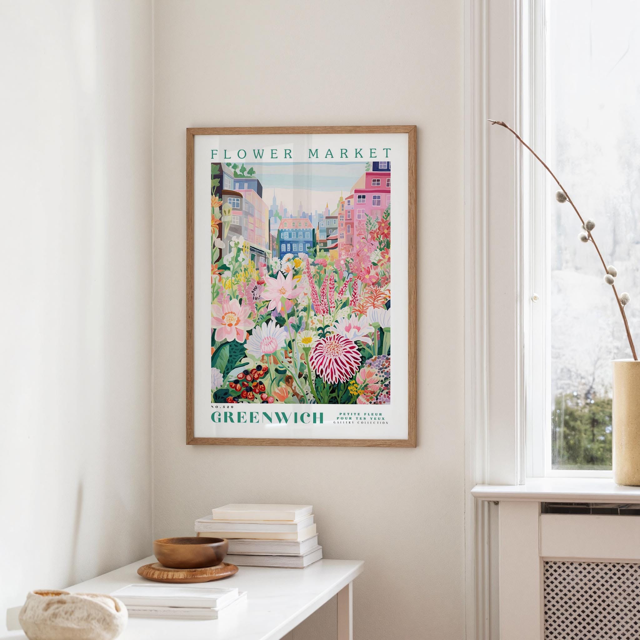 a framed poster of a colorful garden scene hanging on a wall, with a white table and a vase of flowers in the foreground.