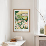 a framed poster of a flower garden hanging on a wall, with a white table and a vase of flowers nearby.