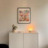 a white cabinet with a potted plant and a lamp on top, and a framed poster of a cityscape hanging on the wall above it.