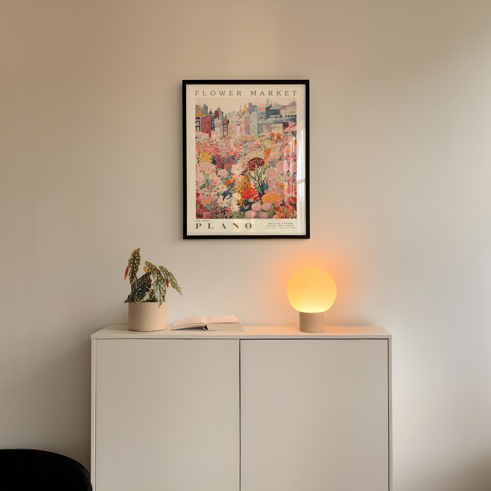a white cabinet with a potted plant and a lamp on top, and a framed poster of a cityscape hanging on the wall above it.