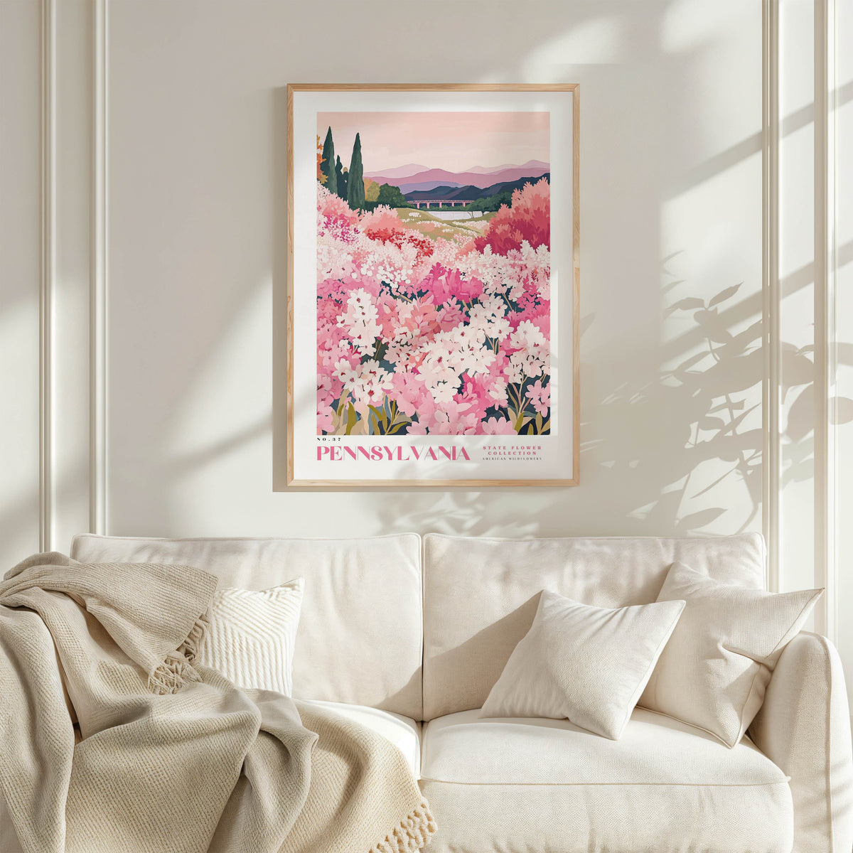 a framed poster of a field of pink flowers hanging on a wall above a white couch with a blanket draped over it.