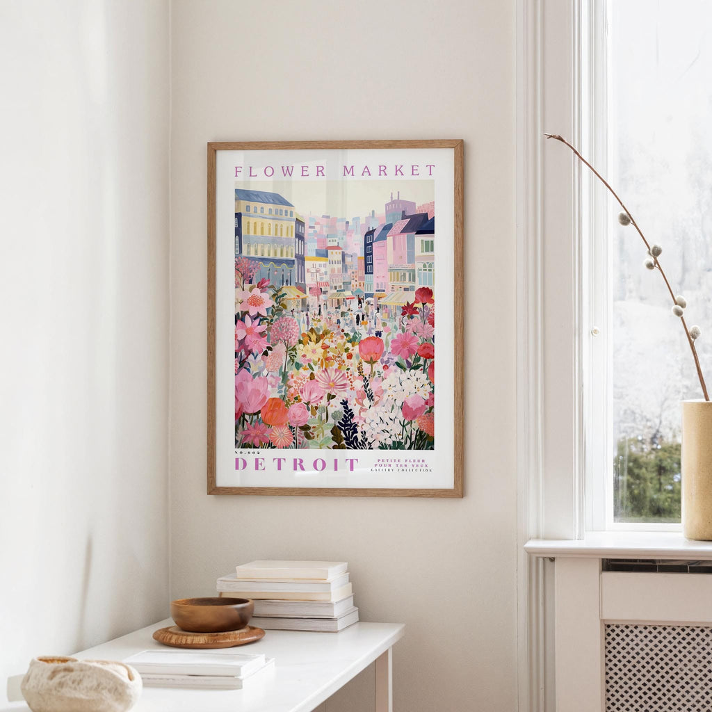a framed poster of a colorful cityscape with flowers hanging on a wall, next to a window and a small table with books and a bowl.
