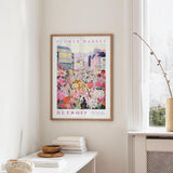 a framed poster of a colorful cityscape with flowers hanging on a wall, next to a window and a small table with books and a bowl.