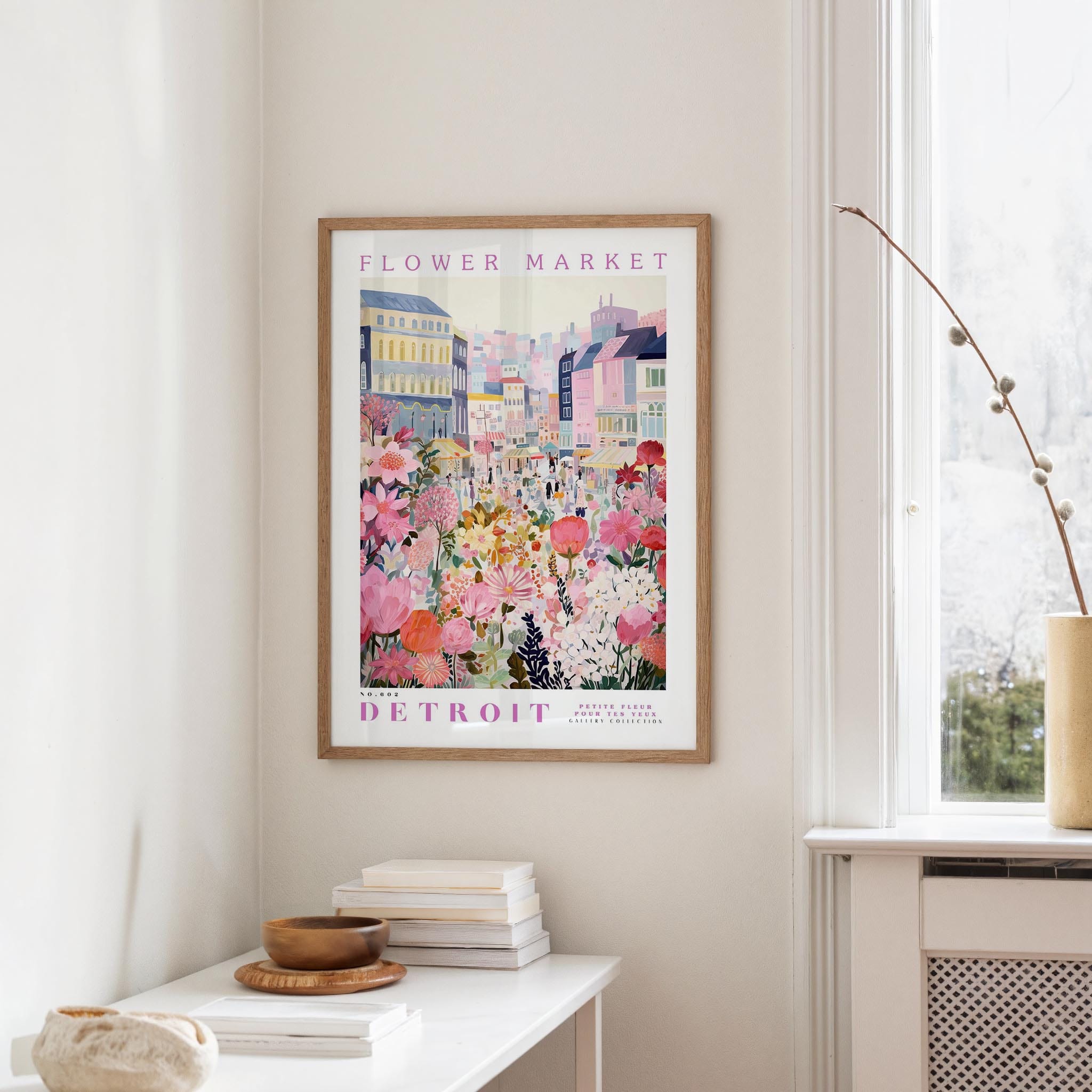 a framed poster of a colorful cityscape with flowers hanging on a wall, next to a window and a small table with books and a bowl.