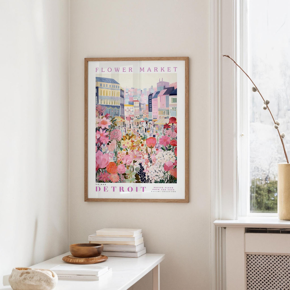 a framed poster of a colorful cityscape with flowers hanging on a wall, next to a window and a small table with books and a bowl.