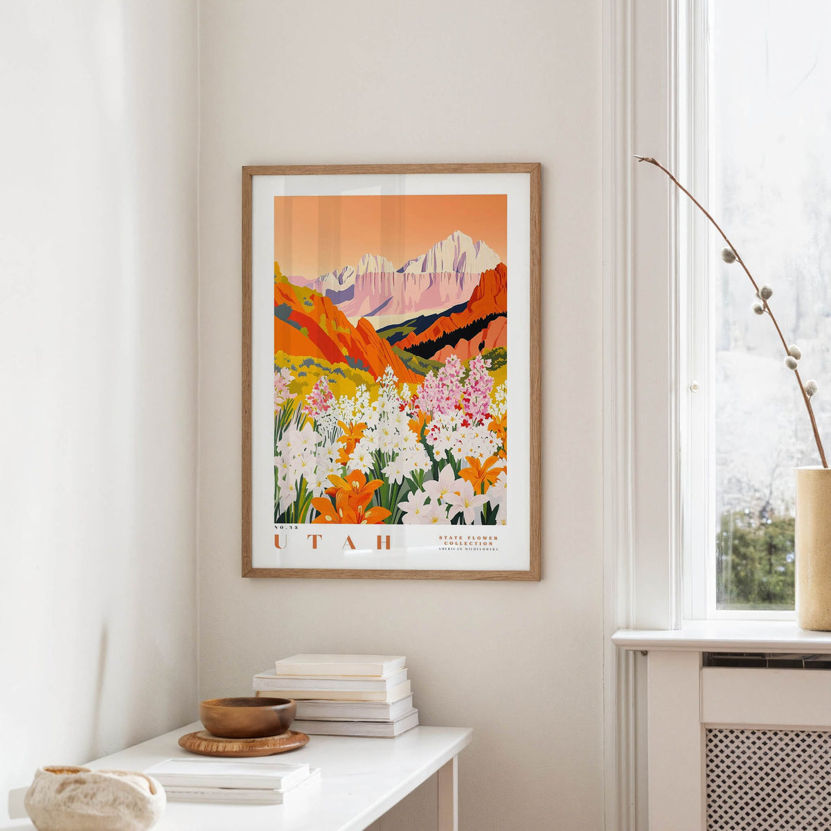 a framed poster of a colorful mountain landscape with flowers hanging on a wall in a room with a white table and a window.