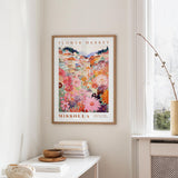a framed poster of a colorful floral scene hanging on a wall, with a white table and a vase of flowers in the foreground.