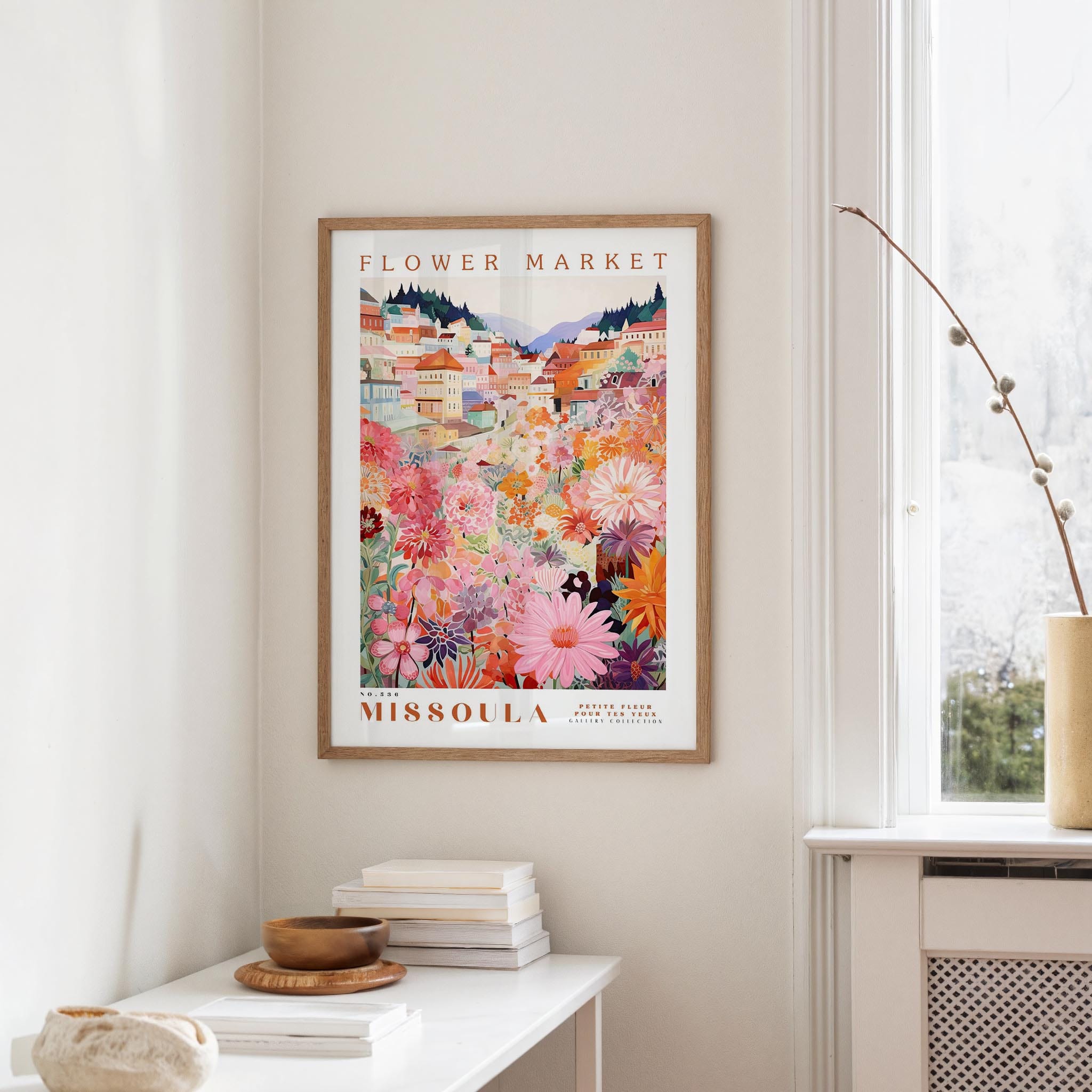 a framed poster of a colorful floral scene hanging on a wall, with a white table and a vase of flowers in the foreground.