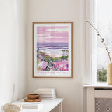 a framed poster of a beach scene with a colorful, abstract landscape, hanging on a wall in a room with a white table and a window.