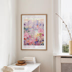 a framed poster of a cityscape with a floral design, hanging on a wall in a room with a window and a small table with books and a vase.