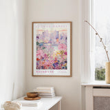 a framed poster of a cityscape with a floral design, hanging on a wall in a room with a window and a small table with books and a vase.