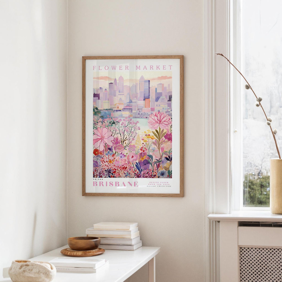a framed poster of a cityscape with a floral design, hanging on a wall in a room with a window and a small table with books and a vase.