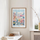 a framed poster of a colorful floral scene hanging on a wall, with a window and a small table with books and a vase in the foreground.