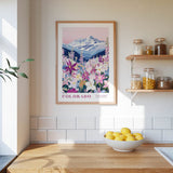a framed poster of a mountain landscape with a variety of flowers, hanging on a wall above a wooden countertop with a bowl of lemons.