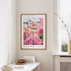 a framed poster of a colorful garden scene, with a cityscape in the background, hanging on a wall in a room.