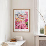 a framed poster of a colorful garden scene, with a cityscape in the background, hanging on a wall in a room.