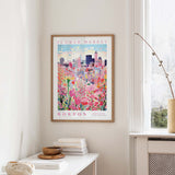 a framed poster of a cityscape with a flower market in the background, hanging on a wall in a room with a window and a table with books and a vase.