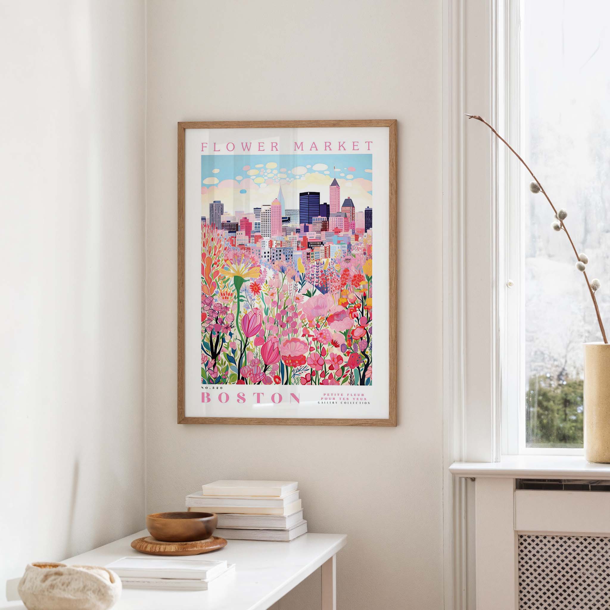 a framed poster of a cityscape with a flower market in the background, hanging on a wall in a room with a window and a table with books and a vase.