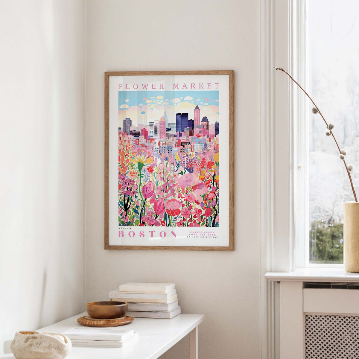 a framed poster of a cityscape with a flower market in the background, hanging on a wall in a room with a window and a table with books and a vase.
