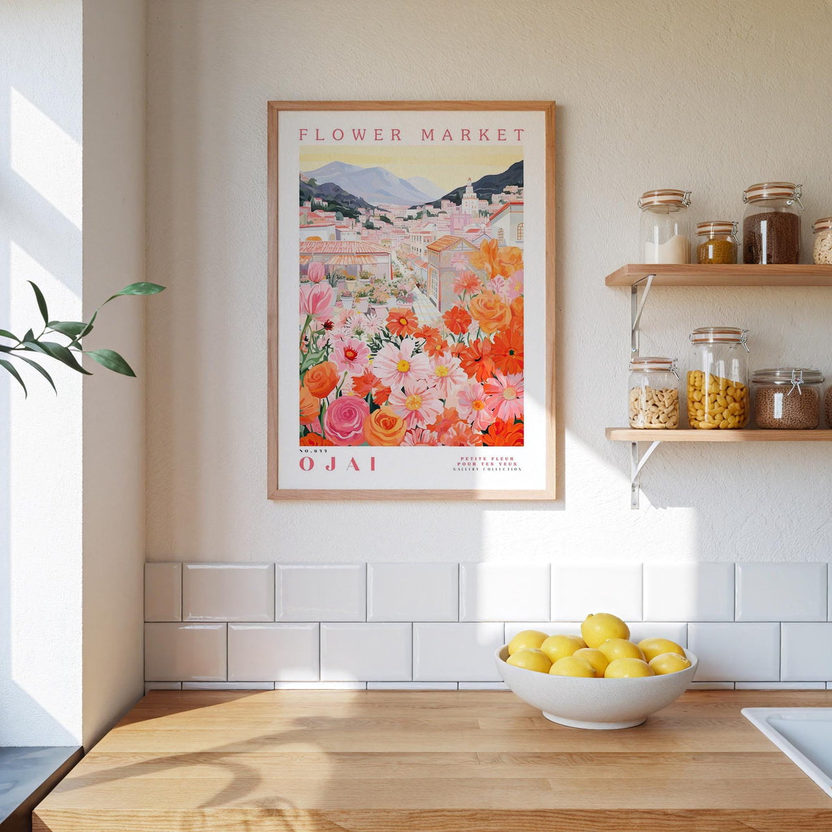 a kitchen scene with a framed poster of a flower market in Ojai, Italy, hanging on the wall above a wooden counter. The poster displays a vibrant illustration of a flower market with colorful flowers and a picturesque town in the background. On the counter, there is a bowl of lemons and a plant, adding a touch of greenery to the scene.