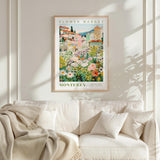 a framed poster of a flower market in Monterey, California, hanging on a wall above a white couch with a blanket draped over it.