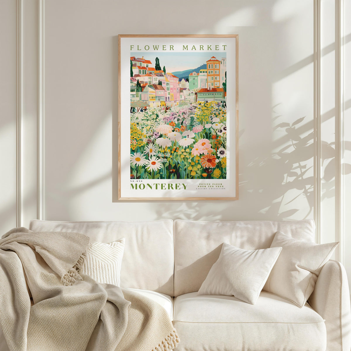 a framed poster of a flower market in Monterey, California, hanging on a wall above a white couch with a blanket draped over it.