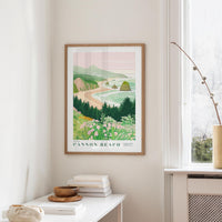 a framed poster of Cannon Beach, a coastal town in Oregon, hanging on a wall in a room with a white table and a vase of flowers.
