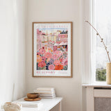 a framed poster of a colorful floral scene hanging on a wall, with a white table and a vase of flowers in the foreground.