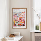 a framed poster of a colorful floral scene hanging on a wall, with a white table and a vase of flowers nearby.