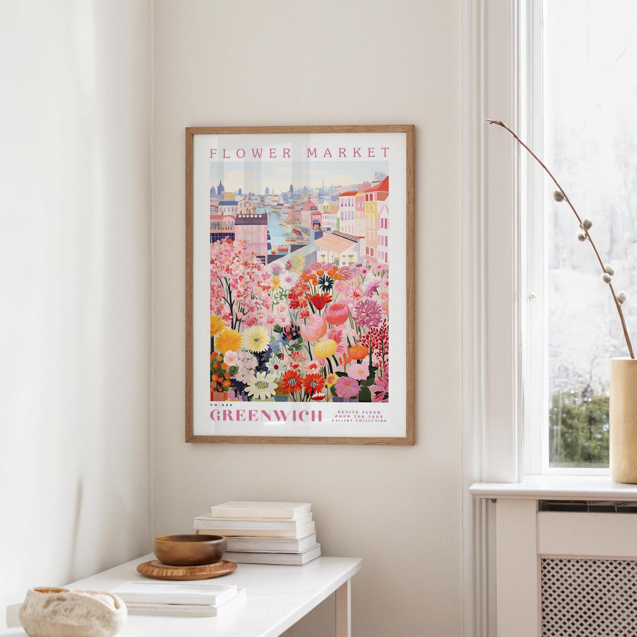 a framed poster of a colorful floral scene hanging on a wall, with a white table and a vase of flowers nearby.