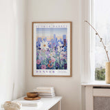 a framed poster of a cityscape with a field of flowers in the foreground, hanging on a wall in a room with a window and a small table with books and a vase.