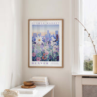 a framed poster of a cityscape with a field of flowers in the foreground, hanging on a wall in a room with a window and a small table with books and a vase.