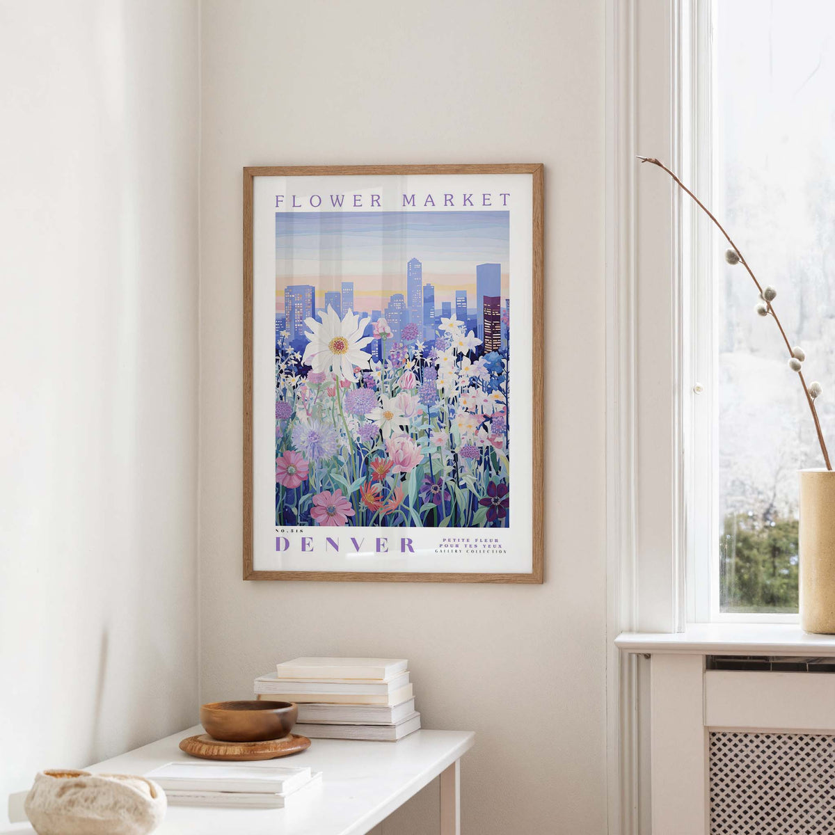 a framed poster of a cityscape with a field of flowers in the foreground, hanging on a wall in a room with a window and a small table with books and a vase.