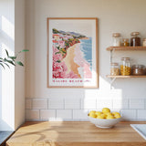 a framed poster of a beach scene with a wooden frame, hanging on a wall above a kitchen counter with a bowl of lemons and jars of food on the counter.