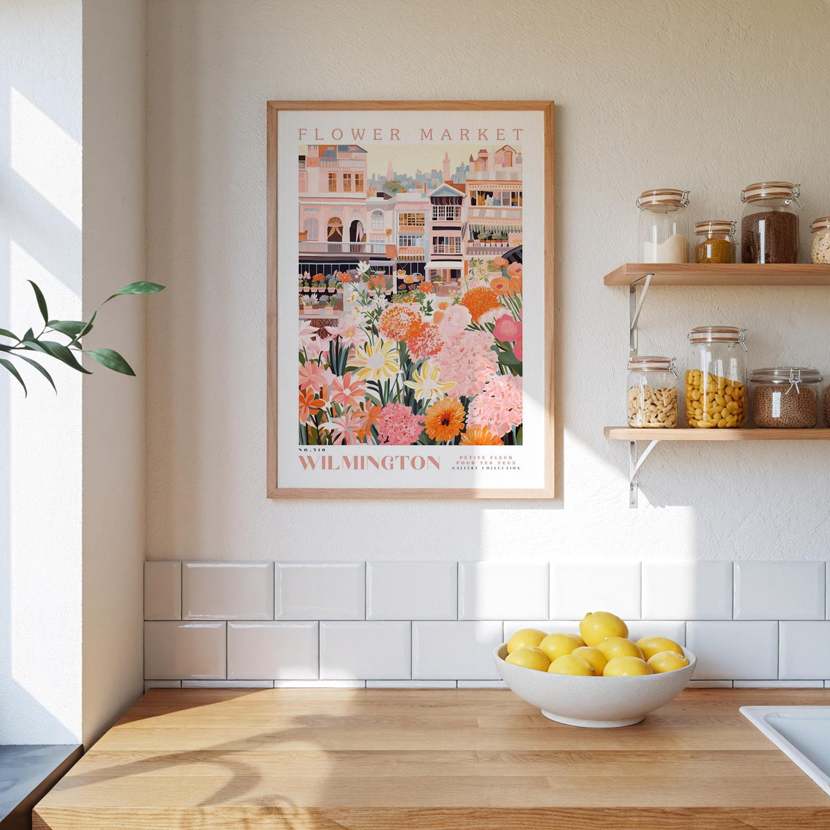 a kitchen scene with a framed poster of a flower market in Wilmington, Australia, hanging on the wall above a wooden counter. The poster displays a variety of colorful flowers and plants, creating a vibrant and lively atmosphere. On the counter, there is a bowl of lemons, adding a touch of freshness to the scene. The overall composition of the image is visually appealing and showcases the beauty of the flower market in Wilmington.