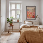 a cozy bedroom with a large bed, a wooden nightstand, and a large window with a plant on the sill. The room has a warm, inviting atmosphere with a neutral color palette and natural elements like the potted plant and the framed poster on the wall.