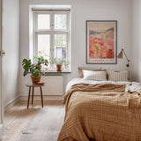 a cozy bedroom with a large bed, a wooden nightstand, and a large window with a plant on the sill. The room has a warm, inviting atmosphere with a neutral color palette and natural elements like the potted plant and the framed poster on the wall.